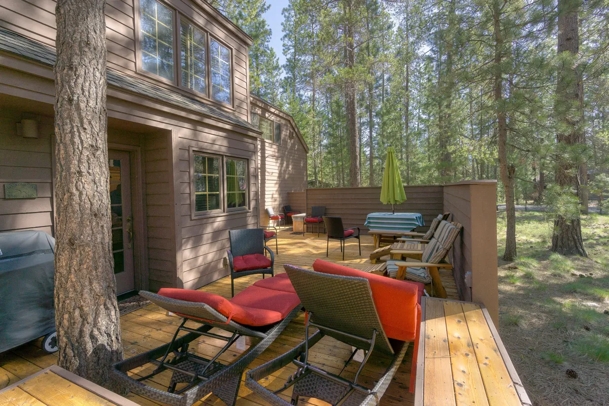 This image features a spacious wooden deck surrounded by tall trees, with comfortable lounge chairs, a dining table, and a large green umbrella creating an inviting atmosphere for outdoor relaxation and dining.