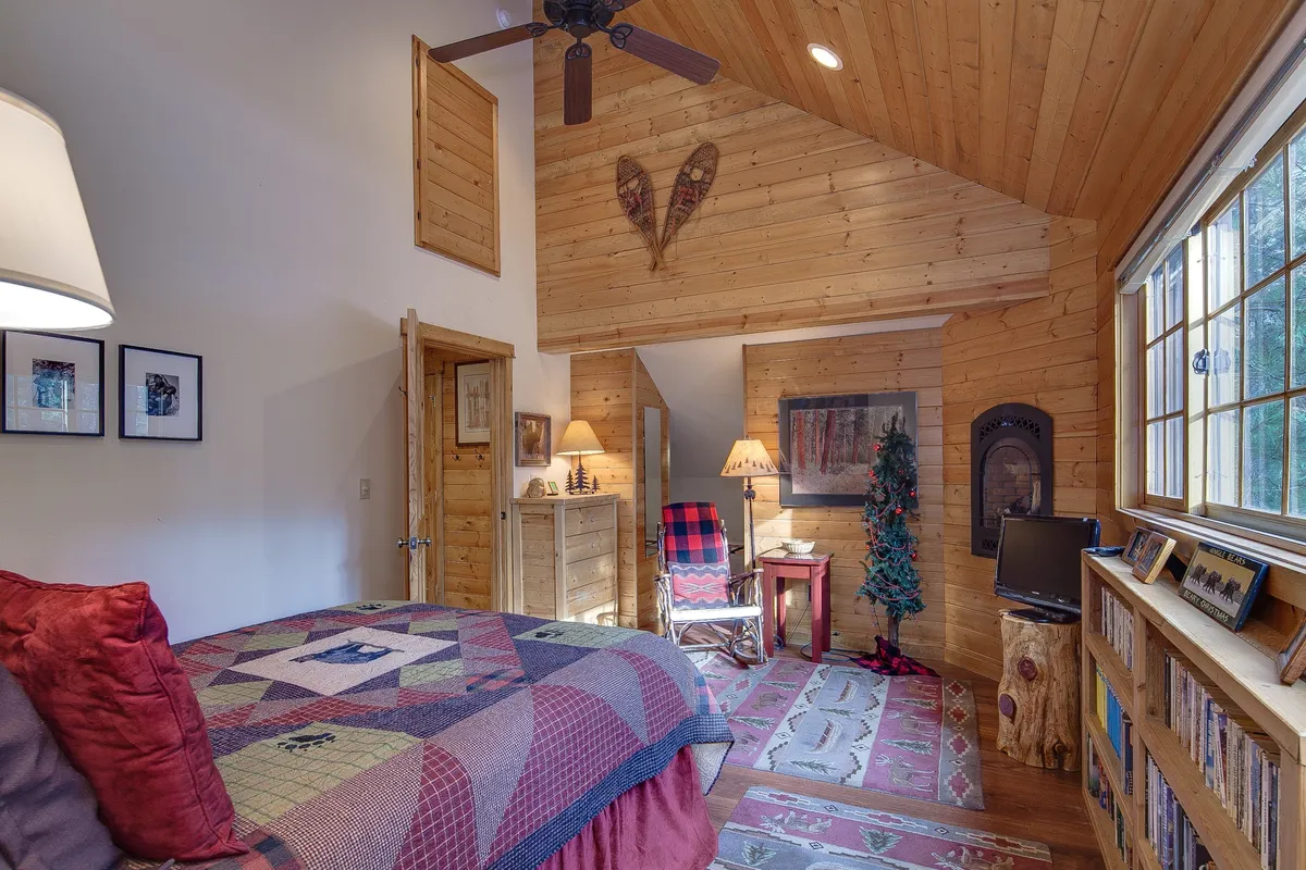 A serene and cozy loft bedroom with a queen bed draped in a quilt, wooden furniture, decorative wall art, a small Christmas tree, and a bookshelf. The peaceful atmosphere underscores the promise of privacy and comfort in the second loft.