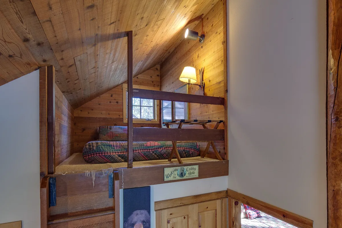 A loft sleeping nook with a wooden ceiling, a cozy loft bed dressed in plaid bedding, a lamp, and soft lighting. The space feels secluded and intimate, perfect for those seeking extra privacy or a whimsical hideaway.