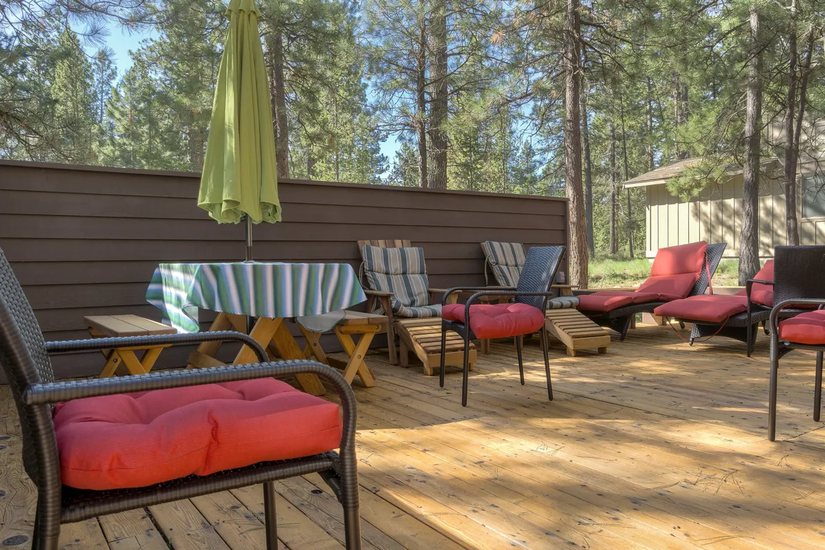 This image captures a wooden deck furnished with red cushioned chairs, a striped tablecloth, and a green umbrella, creating an inviting spot for outdoor dining, casual reading, or socializing. The deck is surrounded by greenery and natural light.