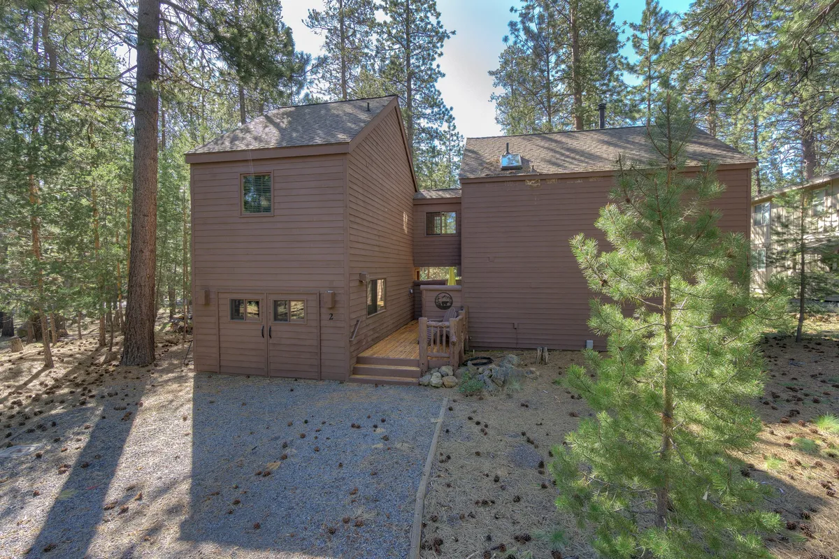 This exterior image features the wooden cabin nestled among tall trees, with a gravel path leading up to the entrance. The tranquil scene is accented by natural landscaping, evoking a sense of peaceful seclusion but also accessibility to outdoor adventure.