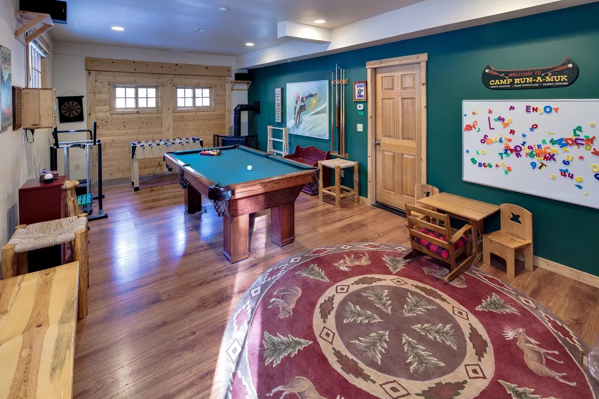 The image showcases a cozy, lodge-inspired living space centered around a pool table, with warm wooden accents and inviting furniture. Playful touches like a whiteboard with colorful letters and a patterned rug hint at family-friendly fun and creative activities. The setting exudes warmth, comfort, and a sense of togetherness, perfect for shared games and laughter.