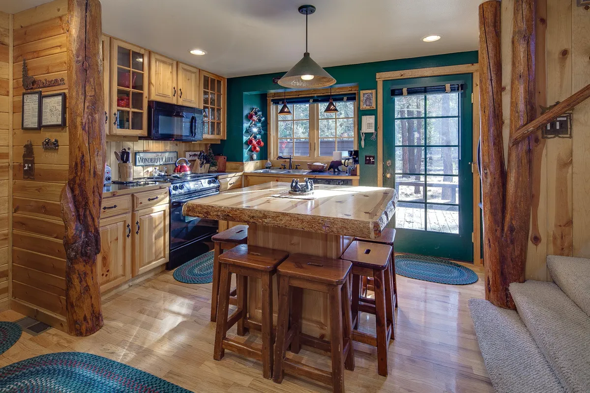 A bright and welcoming kitchen scene featuring a large island with wooden stools, a green accent wall, and expansive windows letting in natural light. The space is staged for group cooking and casual gatherings, aligning with the kitchen’s sociable purpose.