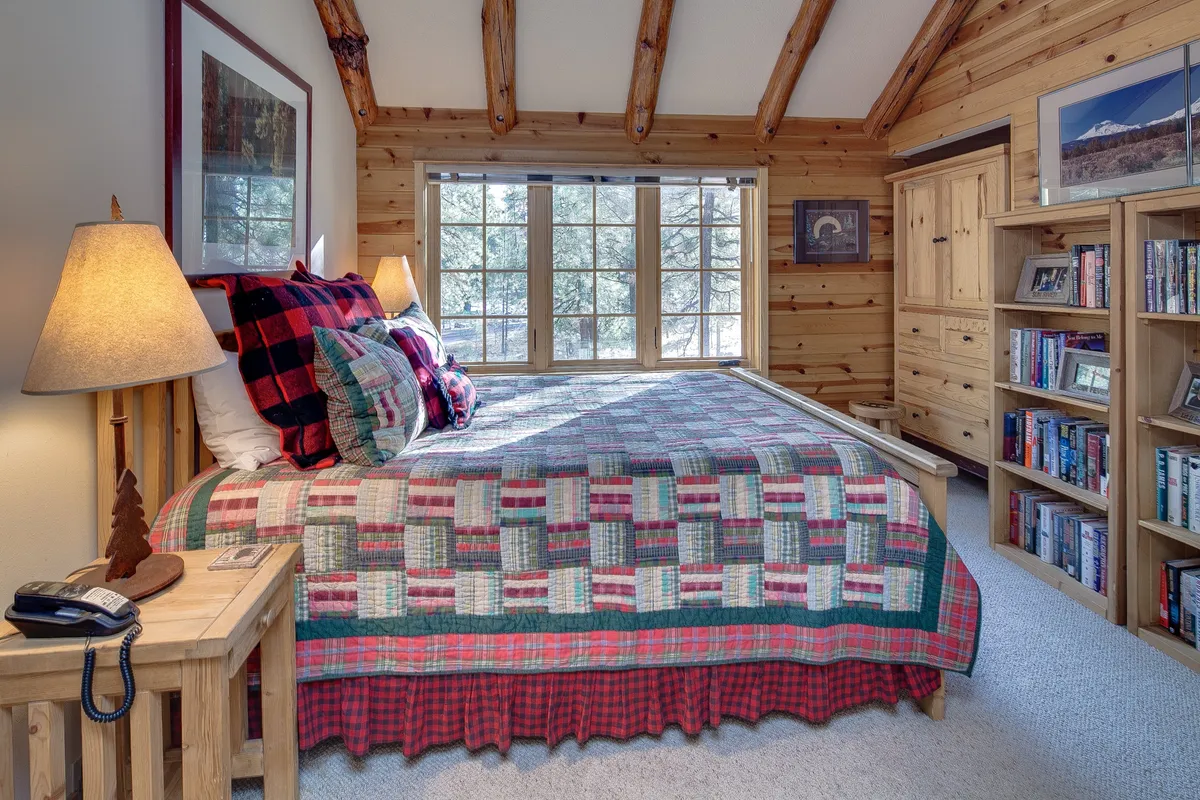 A serene bedroom with a comfortable bed neatly made with a quilt, large windows that let in soft natural light, wooden furniture, and a bookshelf. The space feels calm and restful, ideal for relaxation or reading in privacy.